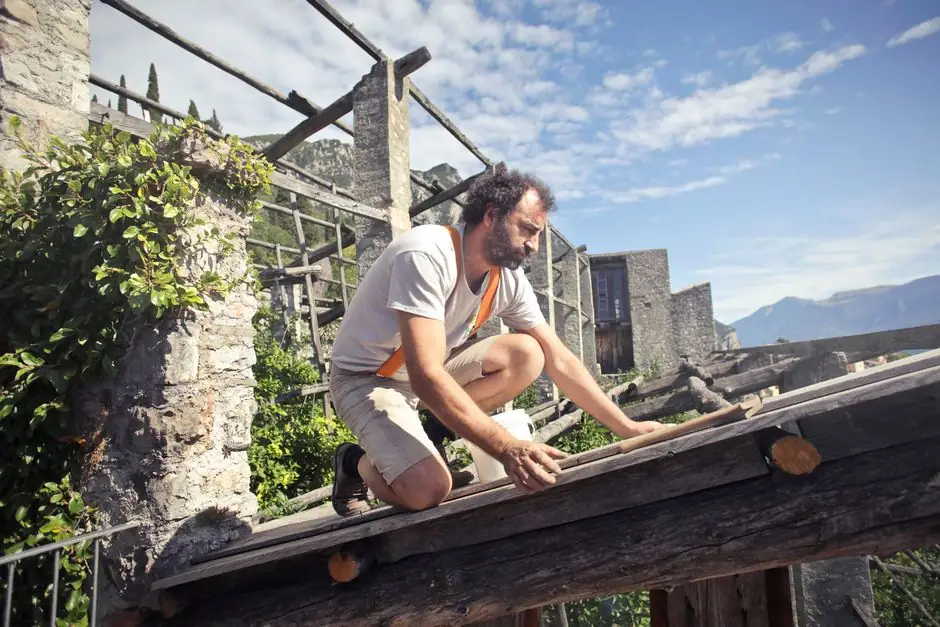 Side view of male builder in summer wear reconstructing rooftop and sitting on wooden beams of aged building on background of scenic landscape in sunny day